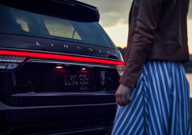 A person is shown near the rear of a 2024 Lincoln Aviator® SUV as the Lincoln Embrace illuminates the rear lights | Cavalier Lincoln in Chesapeake VA