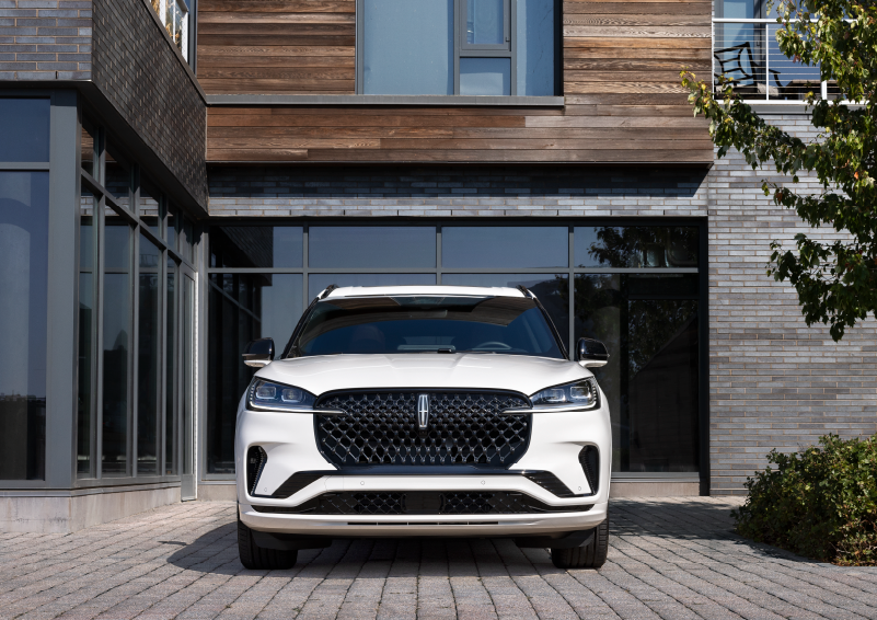 A white 2025 Lincoln Aviator® SUV is shown parked outside a modern building with gray brick and wood accents. | Cavalier Lincoln in Chesapeake VA