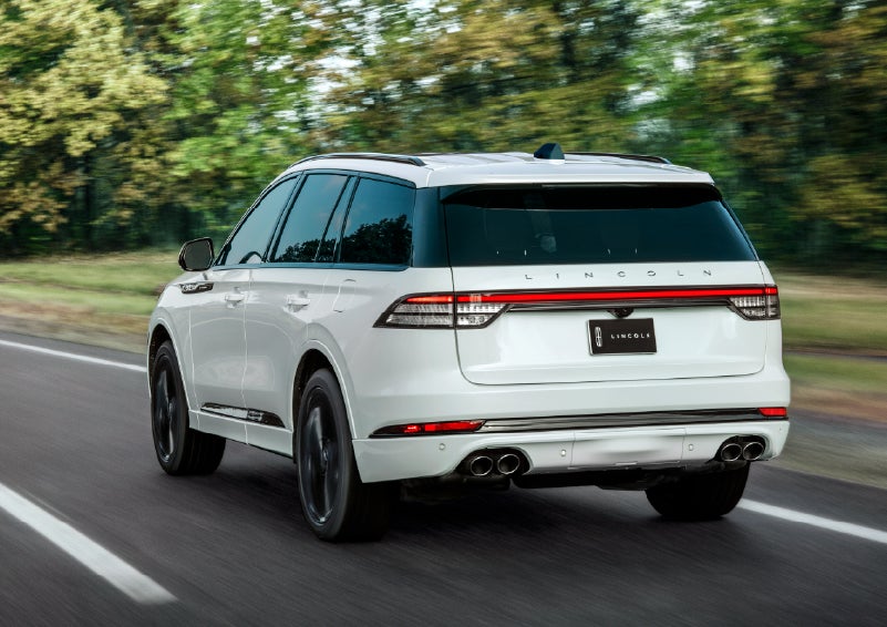 A white 2026 Lincoln Aviator® SUV is being driven away down a road lined with colorful trees. | Cavalier Lincoln in Chesapeake VA