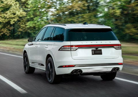 A white 2026 Lincoln Aviator® SUV is being driven away down a road lined with colorful trees. | Cavalier Lincoln in Chesapeake VA